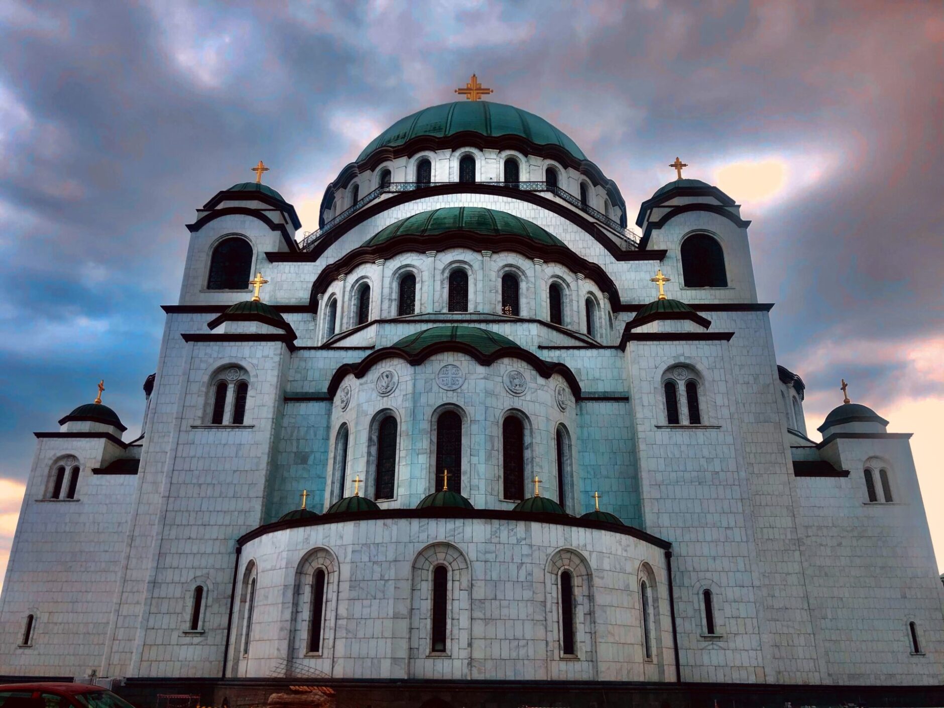 White brick facade and green dome roof of Saint Sava Church against a pink and blue sky 